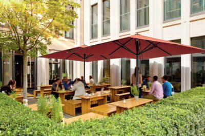 Foto einer gut besuchten Außenterrasse vor einem modernen Bürogebäude, beschattet von zwei großen, roten Sonnenschirmen.