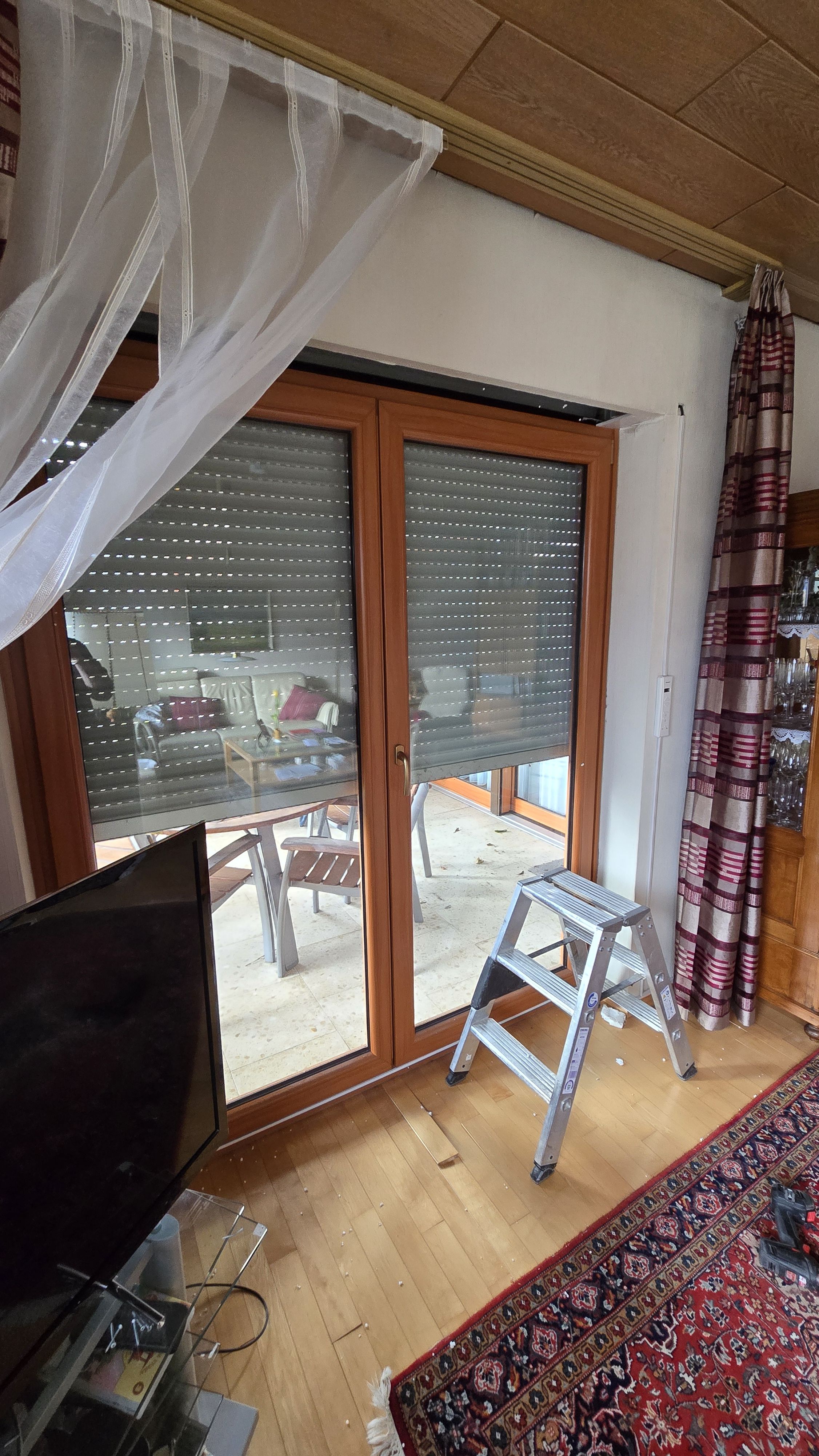 Reparatur eines Rollladens an einer großen Terrassentür mit Holzrahmen; der Behang ist halb heruntergelassen und eine Trittleiter steht für Montagearbeiten bereit.